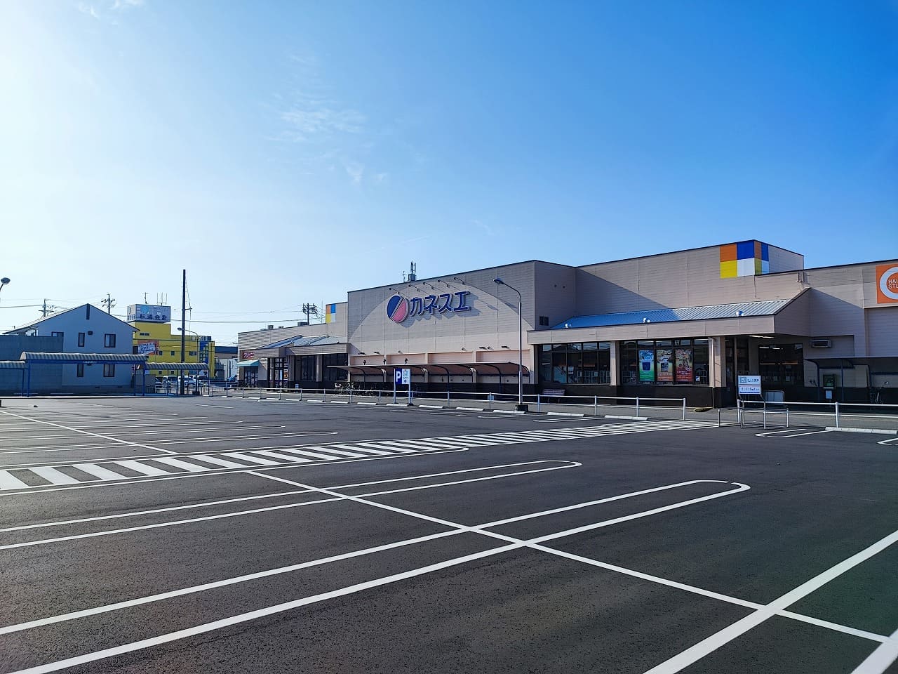 カネスエ国府宮店の店舗の外観と大型駐車場。モダンなグレーの外壁の建物にカネスエのロゴが掲げられ、広大な駐車場には白線で区画整理された多数の駐車スペースが整備されている。青空の下、清潔で利用しやすい商業施設の様子。