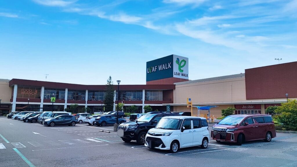 リーフウォーク稲沢の正面外観。緑色の葉っぱロゴが特徴的なLEAF WALKの看板が建物上部に設置されている。2階建ての茶色とベージュの外観で、1階にはガラス張りの店舗が並ぶ。駐車場にはファミリー向けのミニバンや軽自動車、SUVなど様々な車種が駐車しており、車椅子マークのあるバリアフリー駐車場も確認できる。