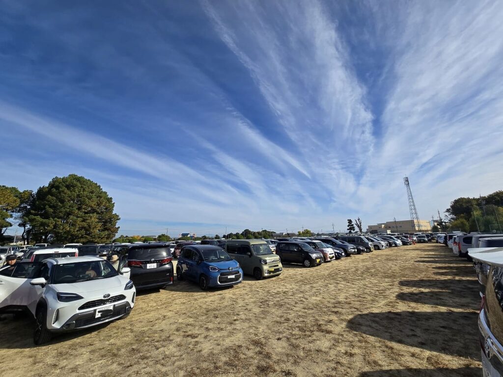 【キャプション】 ``` 西島グラウンドの臨時駐車場 ``` --- 【代替テキスト】 ``` 祖父江イチョウ黄葉まつりの臨時駐車場として開放されている西島グラウンドの様子。広い敷地に多数の車が整然と駐車されており、朝9時前でもすでに多くの来場者が訪れていることがわかる。青空には白い雲が筋状に広がり、秋晴れの良い天気。背景には木々や建物、電波塔が見える。祭り期間中の駐車場の混雑状況を表した写真で、早めの来場が推奨されることを示している。 ```