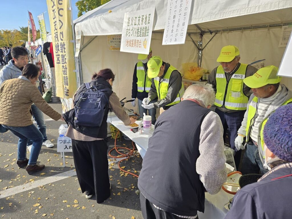 祖父江イチョウ黄葉まつりの銀杏試食コーナーで、黄色い蛍光ベストを着たスタッフが銀杏を調理している様子。白いテーブルの手前では銀杏を揚げる作業が、奥では焼く作業が行われており、来場者が順番に試食を受け取っている。テント上部には「祖父江支所職員まちづくりの会連絡会(地域振興会)」の看板が掲げられている。複数の来場者がテーブルを囲んで試食を待っており、地元のボランティアスタッフによる手作りのおもてなしが感じられる温かい光景。