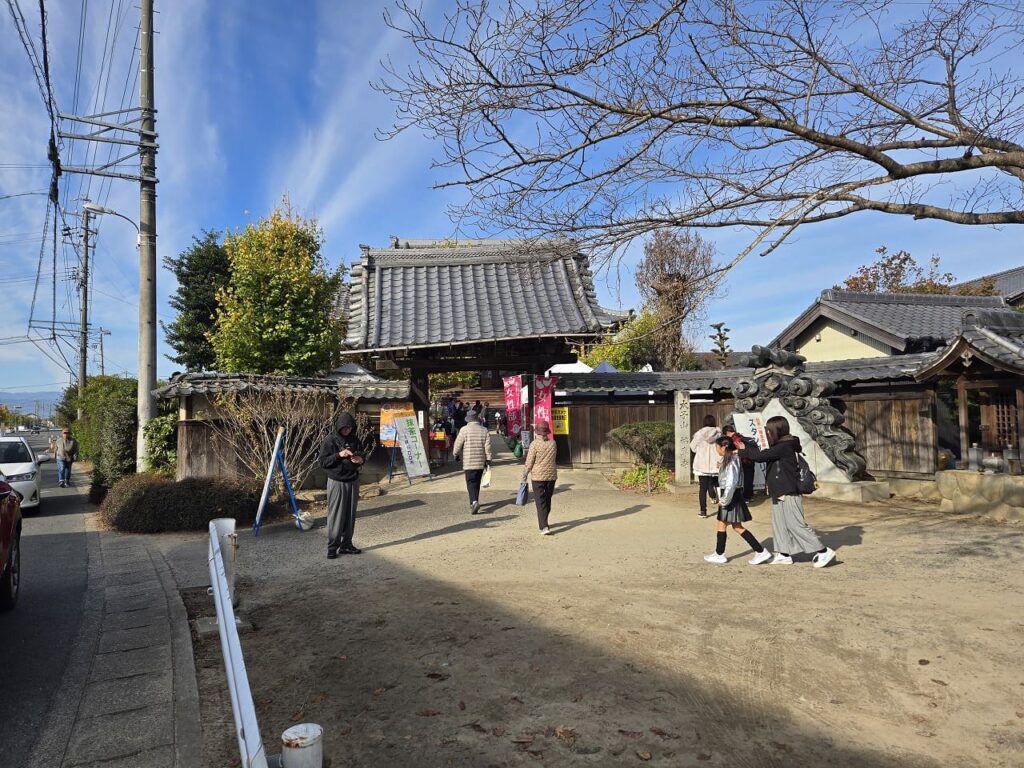 【キャプション】 ``` 祐専寺会場の入り口付近の様子 ``` --- 【代替テキスト】 ``` 祖父江イチョウ黄葉まつりの祐専寺会場入り口付近を撮影した写真。伝統的な日本建築の本堂と瓦屋根が見え、入り口には赤いのぼりが立てられている。晴天の青空の下、境内には複数の来場者が歩いており、左側には黄色く色づいたイチョウの木が見える。道路沿いには電柱と車が停まっており、地域に根ざした歴史あるお寺の雰囲気が伝わる会場の風景。 ```