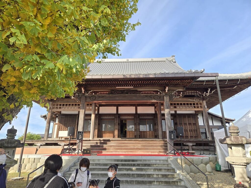 祐専寺会場の本堂を正面から撮影した写真。伝統的な日本建築の本堂は精巧な木彫りの装飾が施され、瓦屋根と格子戸が美しい。階段の前には赤い絨毯が敷かれており、ステージイベントの準備がされている。左側には黄色く色づいたイチョウの大木が本堂を彩り、晴天の青空とのコントラストが美しい。手前には来場者が数名いて、これからのイベントを待っている様子。祐専寺会場のメインステージとなる場所。