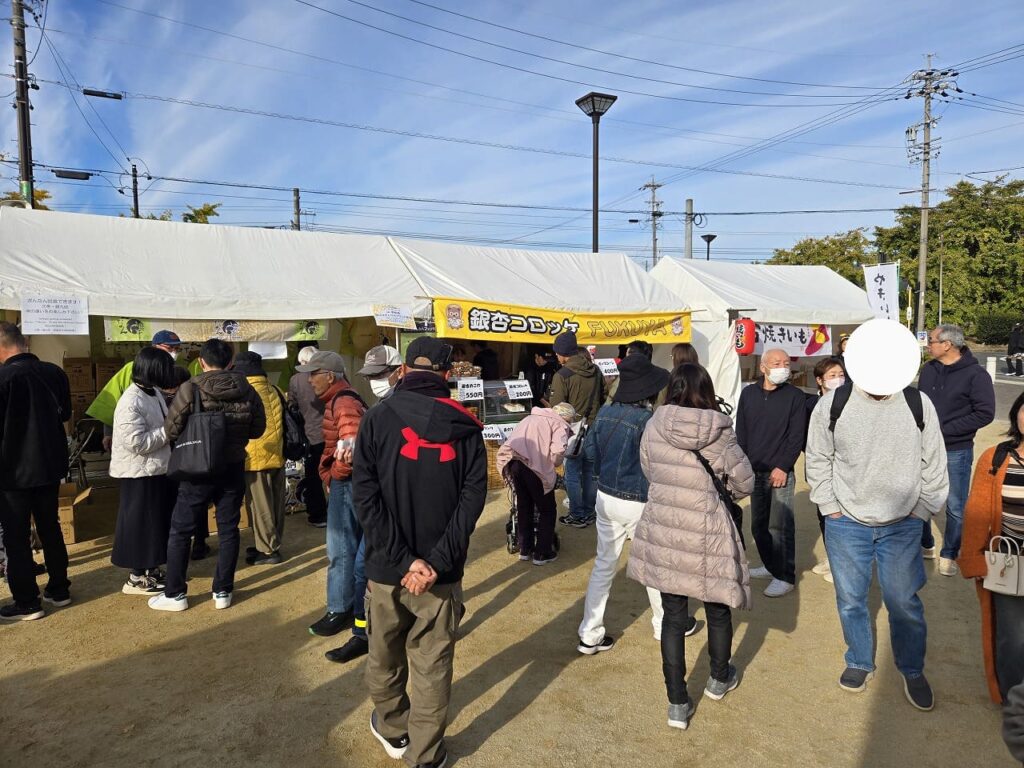 祖父江イチョウ黄葉まつり会場の祖父江ぎんなんパークで、複数のテントが立ち並び多くの来場者で賑わっている様子。中央のテントには「銀杏コロッケ」の黄色い看板が掲げられており、その前には長い行列ができている。晴天の青空の下、家族連れやカップルなど様々な年齢層の人々が会場を歩いている。白いテントが3つ並び、屋台や物産販売が行われている祭りの活気ある風景。