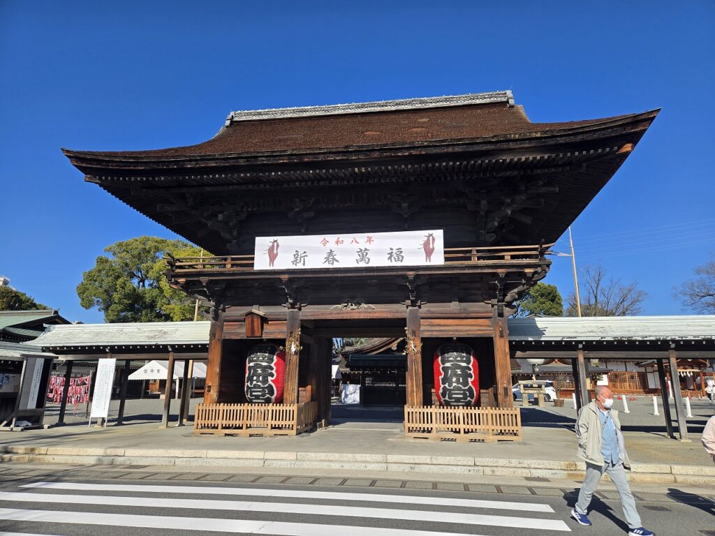 国府宮神社の楼門を正面から撮影した写真。青空を背景に、重厚な木造の楼門がそびえ立つ。「令和八年 新春萬福」と書かれた看板が掲げられ、左右には「国府宮」と書かれた大きな赤い提灯が飾られている。正月を迎える準備が整った様子