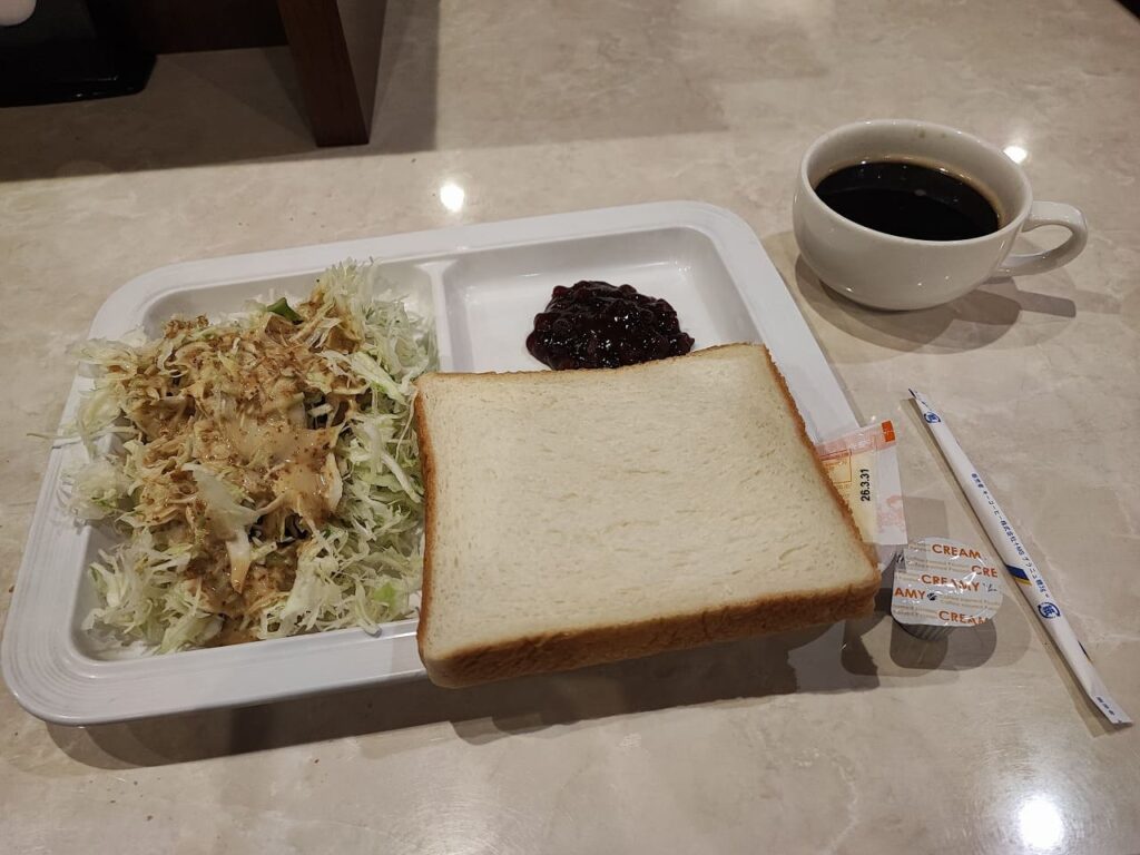 楽だの湯稲沢店の平日限定無料モーニングセットの写真。白い長方形のトレーに食事が盛り付けられており、左側にはドレッシングがかけられた千切りキャベツのサラダ、中央には食パン1枚とその上に小倉あん(またはジャム)が添えられている。食パンの横には小さなマーガリンのカップとプラスチックナイフが置かれている。トレーの右側には白いコーヒーカップに入ったホットコーヒーが添えられている。このモーニングセットは平日(月曜から金曜)の朝7時30分から10時まで提供され、入浴料550円のみで無料で利用できるサービス。パンに付けるものは6種類から2種類選ぶことができ、コーヒーはおかわり自由となっている。大理石調のテーブルの上に配置され、施設内の食事処で提供されている様子