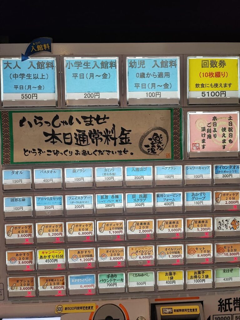 楽だの湯稲沢店の入館料金を示す券売機の写真。上部には料金表示があり、大人(中学生以上)平日550円、小学生平日200円、幼児(0歳から適用)平日100円、回数券10枚綴り5100円(飲食にも使えます)と記載されている。中央には「いらっしゃいませ 本日通常料金」と書かれた案内板があり、その下に多数の券売機ボタンが並んでいる。タオル150円、バスタオル400円、歯ブラシ100円、カミソリ100円、入浴タオル200円などのアメニティ類、固形石鹸100円、アロマ点セット350円などの入浴用品、ボディケア各種(10分単位で価格設定)、あかすり、オイルリンパ、キャンペーンメニューなど多様なサービスボタンが配置されている。一部のボタンには赤いバツ印がついており、売り切れまたは利用不可を示している。券売機は現金のみ対応で、新500円硬貨と新紙幣が使用可能と表示されている