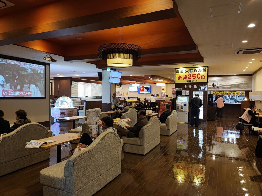 楽だの湯稲沢店の食事処と休憩エリアの全景写真。手前には大型テレビスクリーンが設置されたソファ席があり、複数の利用客が新聞や雑誌を読みながらくつろいでいる。天井には和風の間接照明が施され、暖かみのあるオレンジ色の光で空間を演出している。中央から奥にかけては食事エリアが広がっており、テーブル席やカウンター席が配置されている。奥の壁には黄色い看板で「毎週 火曜めん類 全品250円 開催中!!」と大きく書かれており、火曜日のサービスデーを告知している。食事カウンターには複数の利用客が並んでおり、食券を購入して食事を注文している様子が見える。床は光沢のある茶色のフローリングで清潔感があり、全体的に落ち着いた雰囲気の空間となっている。天井にはエアコンや埋め込み型の照明が設置され、快適な環境が整備されている。入浴後の食事や休憩に適した広々とした空間で、一日中滞在できる設備が充実している