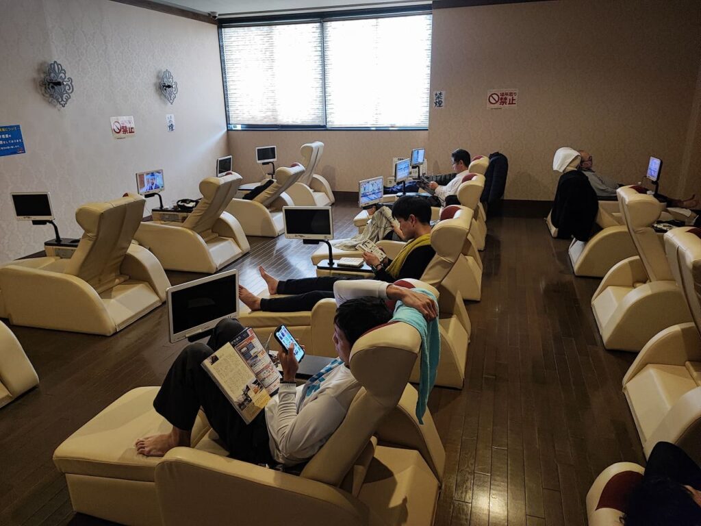 楽だの湯稲沢店のリラックスルームの写真。広々とした室内にベージュ色の電動リクライニングシートが複数台並んでおり、各シートには個別のテレビモニターが設置されている。利用客が思い思いにくつろいでいる様子が見え、手前の利用客は漫画を読みながらリラクライニングシートに座っている。奥の方では複数の利用客がテレビを見たり、スマートフォンを操作したりしている。床は木目調のフローリングで清潔感がある。壁には禁煙マークが掲示されており、大きな窓からは自然光が差し込んでいる。このエリアには約2000冊の漫画が用意されており、入浴料のみで自由に読むことができる。一日中ゆっくり過ごせる空間となっており、入浴前後の休憩やリラックスに最適な設備が整っている