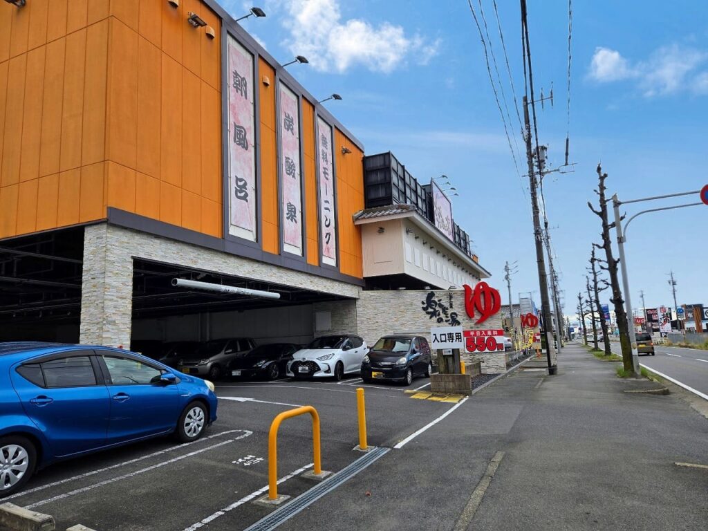 愛知県稲沢市にある楽だの湯稲沢店の外観写真。オレンジ色の壁面が特徴的な2階建ての建物で、建物正面には「飲」「楽」「食べ放題」などと書かれた縦長の看板が複数掲げられている。建物1階部分は駐車場になっており、白や黒の乗用車が数台停まっている。建物前には広い平面駐車場があり、青い車が駐車されている様子が見える。建物の右側には大きく赤い文字で「ゆ」と書かれた看板が掲げられ、その下に「楽だの湯」の店舗看板がある。さらに右側には「入口専用 土日祝日 650円」と書かれた看板が設置されている。青空に白い雲が浮かぶ晴天の日中に撮影された写真で、周辺は郊外の幹線道路沿いの立地であることがわかる。電線や街路樹、他の店舗の看板なども写っており、車でのアクセスが便利な立地であることが伺える