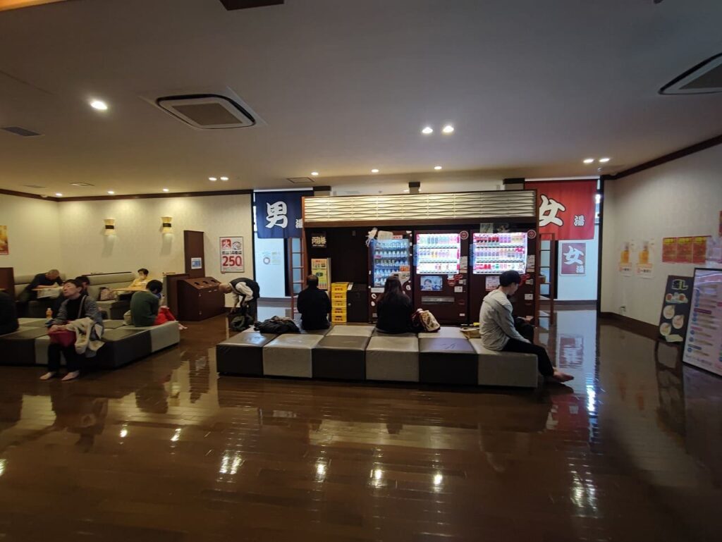 楽だの湯稲沢店の館内休憩スペースの写真。光沢のある茶色の床が敷かれた広々とした空間で、中央には白と灰色のチェック柄の低いソファが複数配置されており、複数の利用客がくつろいでいる様子が見える。奥には男湯と女湯の入口があり、それぞれ紺色の暖簾に「男」、赤色の暖簾に「女」と白い文字で書かれている。暖簾の間には3台の飲料自動販売機が並んでおり、冷たい飲み物を購入できる。左側の壁には「肉々250」と書かれた赤いポスターが掲示されており、火曜日の麺類250円サービスの案内と思われる。天井には埋め込み型の照明とエアコンが設置されている。右側の壁にはメニュー表や案内掲示物が貼られている。全体的に清潔で落ち着いた雰囲気の休憩エリアとなっており、入浴前後にゆっくりできるスペースが確保されている