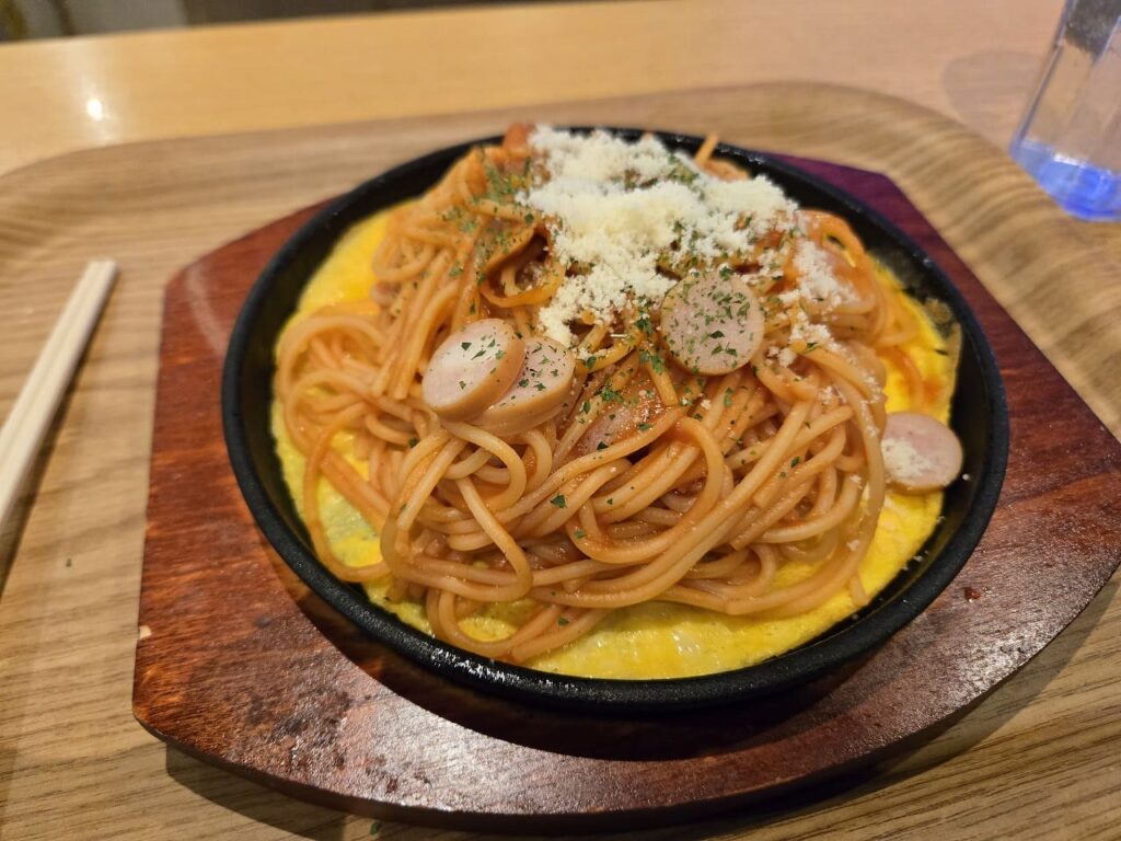 楽だの湯稲沢店で毎週火曜日に250円で提供される鉄板ナポリタンの写真。黒い鉄板の上に黄色い卵が敷かれ、その上にケチャップ味のスパゲッティが盛り付けられている。ナポリタンの上には粉チーズがたっぷりとかけられ、パセリが散らされている。具材としてマッシュルームやタマネギが入っており、湯気が立ち上る熱々の状態で提供されている。鉄板は木製の台座の上に置かれ、左側には割り箸が添えられている。通常価格のナポリタンが火曜日だけ全品250円になる人気のサービスデーメニューで、朝から晩まで一日中この価格で楽しめる。ボリュームもあり、入浴後の食事として多くの利用客に人気の一品。テーブルの上にはグラスに入った飲み物も見える