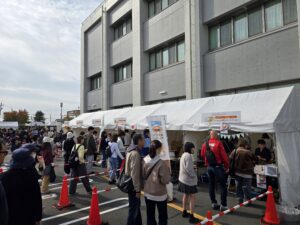 稲沢市役所前の駐車場で開催された稲沢カレーフェスティバル2025の会場。背景には稲沢市役所の灰色のコンクリート建物が見え、その手前に白いテントの出店ブースが複数並んでいる。各ブースの上部にはメニュー看板が掲げられており、Kitahachiなどの店舗名が確認できる。会場には多数の来場者が詰めかけ、赤いカラーコーンと赤白のロープで区切られた動線に沿って行列を作っている。来場者は秋冬の服装で、ベージュやグレー、黒など落ち着いた色合いのコートやジャケットを着用している。右側のテントブースには赤いジャンパーを着たスタッフが対応している様子が見える。曇り空だが明るい日中の雰囲気で、家族連れや若者、中高年など幅広い年齢層の来場者で賑わっている。