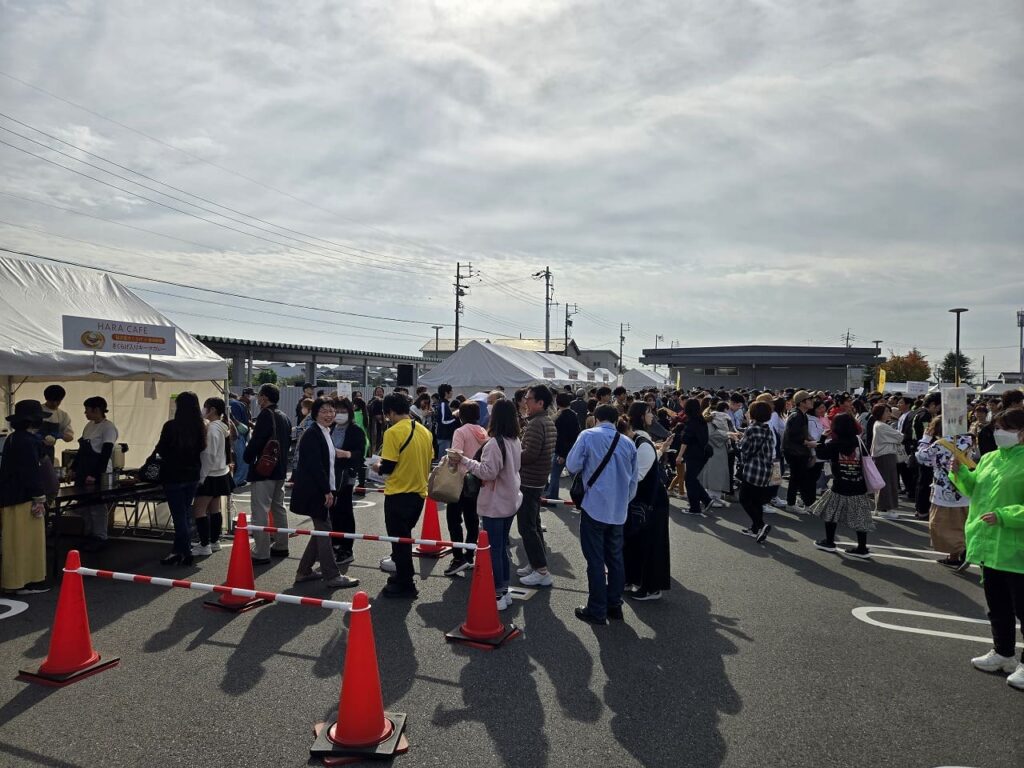 稲沢カレーフェスティバル2025の会場全体の様子。駐車場エリア一帯に大勢の来場者が集まり、各店舗ブースの前には長い行列ができている。手前には赤いカラーコーンと赤白のロープで区切られた動線があり、来場者の流れを整理している。左側には白いテントの店舗ブースが並び、中央から奥にかけて数百人規模の来場者が密集している。黄色やピンク、青などカラフルな服装の来場者が目立ち、家族連れや若者グループなど幅広い年齢層が参加している。背景には住宅街と電柱、曇り空が広がり、秋の穏やかな天候。会場全体が活気に満ちた賑わいを見せている。
