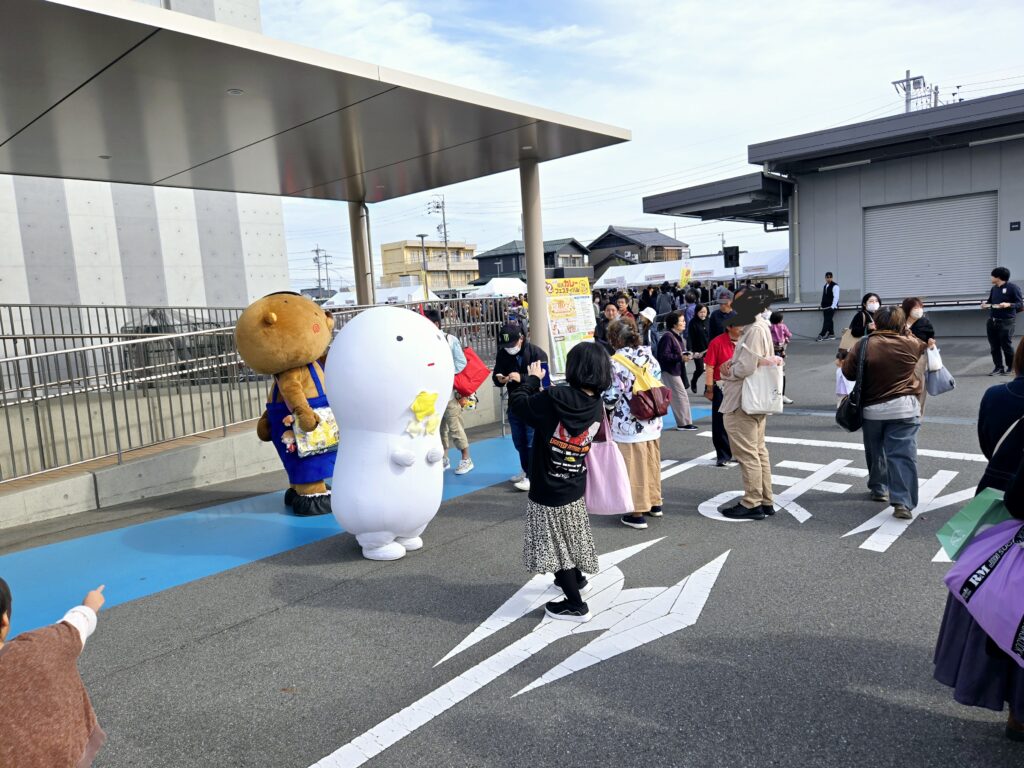 稲沢カレーフェスティバル2025の会場風景。稲沢市役所駐車場で開催され、青空の下、大勢の来場者で賑わっている。手前には2体のゆるキャラが登場。左側は茶色い丸い頭に青い衣装のキャラクター、右側は白い雪だるま型のキャラクター（おそらく稲沢市公式キャラクターのいなッピー）。子どもたちや家族連れがキャラクターと記念撮影をしている。奥には白いテント屋根の下に出店ブースが並び、多くの来場者が行列を作っている。地面にはイベント用の青いカーペットが敷かれ、道路には横断歩道風の白線マーキング。背景には稲沢市役所の建物と周辺の住宅街が見える。秋晴れの良い天候で、ファミリー向けイベントの和やかな雰囲気。