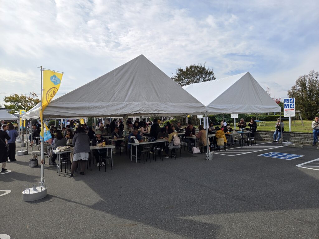 稲沢カレーフェスティバル2025の飲食スペース。大型の白いテント2張りが設置され、その下に長テーブルと椅子が多数並べられている。テント内の座席はほぼ満席で、多くの来場者がカレーを食べている。座席が埋まっているため、テントの外側やその周辺の空きスペースで立ち食いや座り込んで食事をする人々も多く見られる。左手前には黄色いイベント用の旗が立ち、右奥には車椅子マークの青い看板が見える。背景には広い緑地や駐車場、秋の木々が広がり、青空に薄い雲が流れる天候の良い日。会場全体が多くの家族連れや来場者で賑わっている様子。