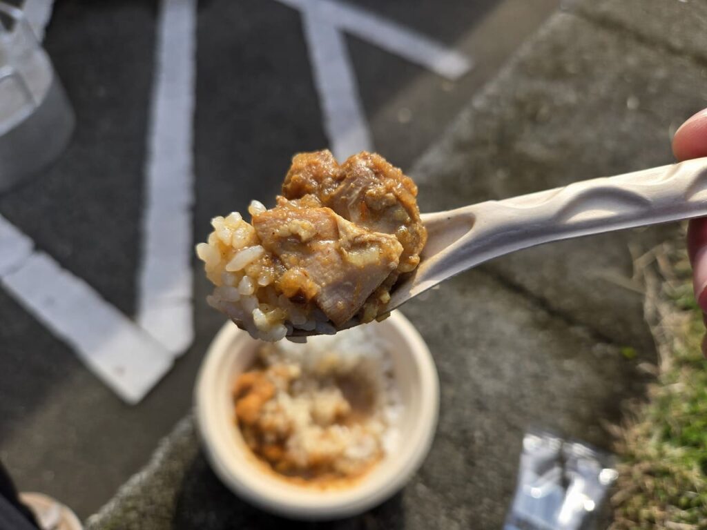 屋外で撮影された料理写真。白いプラスチックスプーンの上に、スパイスで濃いオレンジ色に染まった鶏肉とご飯が一口分すくわれている。鶏肉は角切りで、しっかりとスパイスカレーのルーが絡んでいる。背景には白い紙製の容器に盛られた同じカレーライスが見え、地面はコンクリートで屋外イベント会場の雰囲気。手に持たれたスプーンから湯気は見えないが、食べごろの温かさを感じさせる盛り付け。稲沢カレーフェスティバル2025で提供されたOREGAYAREBAのスパイスチキンカレー。