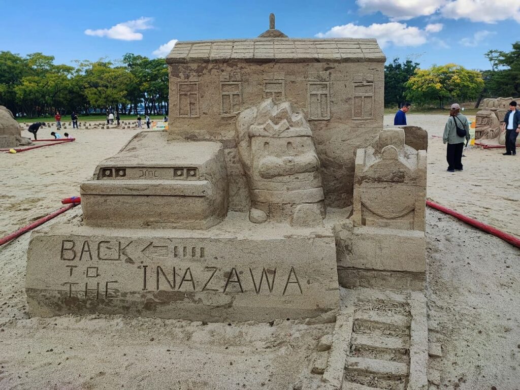 稲沢サンドフェスタ2025の砂像作品「BACK TO THE INAZAWA」。JR貨物稲沢駅開業100周年を記念したテーマで、砂で制作された稲沢駅の建物、貨物列車、機関車が立体的に表現されている。駅舎は瓦屋根と窓が細かく彫刻され、手前には「BACK TO THE INAZAWA」の文字が砂に刻まれている。建物の上部には稲沢市のマスコットキャラクター「イナッピー」が笑顔で彫刻されており、親しみやすい作品になっている。背景には青空と緑の木々が広がり、砂丘エリアで他の来場者が砂像を見学している様子も写っている。作品全体が木曽川の砂を使用した精密な彫刻技術で制作されており、稲沢市の産業の歴史を象徴する作品となっている。