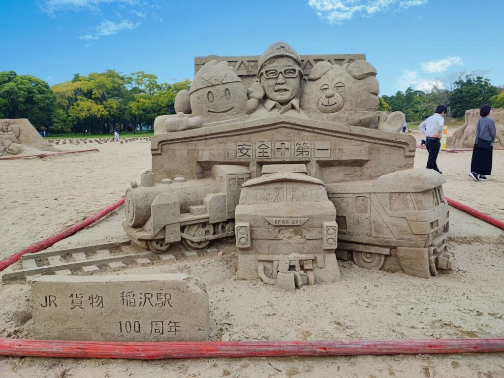 稲沢サンドフェスタ2025の砂像作品「交全志第一（交通安全第一）」。JR貨物稲沢駅開業100周年を記念したテーマで、中央に「交全志第一」と書かれた駅舎の看板があり、上部には稲沢市のマスコットキャラクター「イナッピー」（左側、笑顔のキャラクター）と「ウルフィー」（右側、くまのようなキャラクター）、そして中央にJRの制服を着た駅員が砂で立体的に表現されている。下部には「EF66 27」と記されたSLタイプの機関車と貨物列車、トラックが精密に彫刻されており、手前には「JR貨物 稲沢駅 100周年」の文字が刻まれている。背景には青空と緑の木々が広がり、砂丘エリアで来場者が他の砂像を見学している様子も写っている。作品全体が木曽川の砂を使用した精密な彫刻技術で制作されており、稲沢市の産業と交通安全への願いを表現した作品となっている。