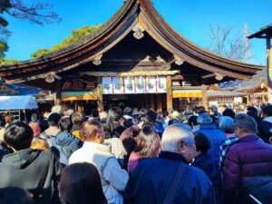 国府宮神社の初詣の様子。青空の下、拝殿前に多数の参拝者が並んでいる。黒い木造の拝殿は大きな屋根が特徴的で、正面には白い注連縄と紙垂が飾られている。参拝者たちは冬服姿で、三が日特有の混雑が見られる。左右には授与所や社務所が配置され、新年を祝う雰囲気が漂う。2024年1月2日撮影。