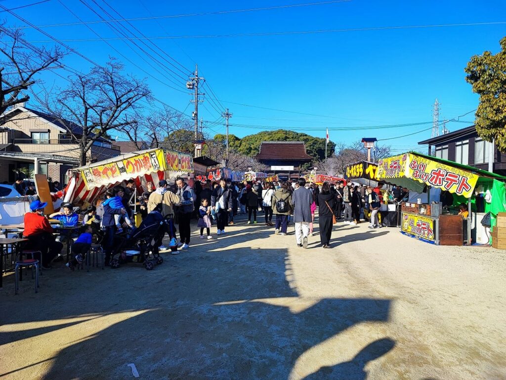 国府宮神社の参道に立ち並ぶ初詣の屋台。快晴の青空の下、参道の両側にカラフルな看板を掲げた屋台が並び、多くの参拝客で賑わっている。左側にはりんご飴やお好み焼きの屋台、右側にはポテトなどの屋台が出店している。参道の奥には神社の建物と緑の木々が見え、周辺には住宅が立ち並ぶ。家族連れやカップルなど様々な世代の参拝客が屋台を楽しむ様子が見られる。2024年1月2日撮影。