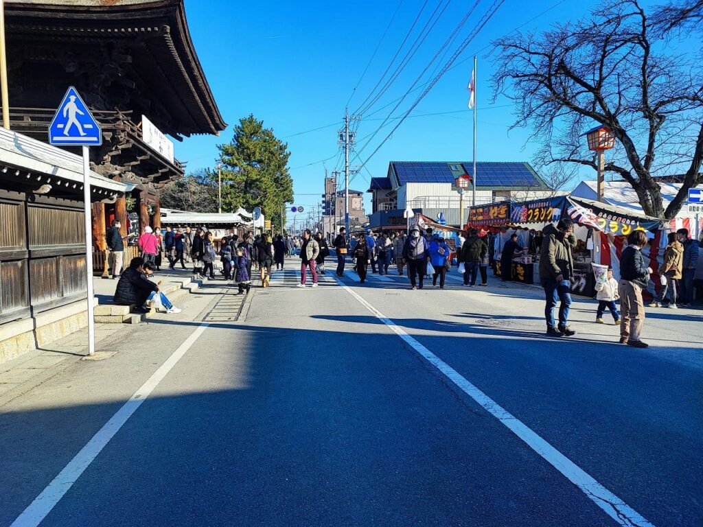 交通規制により歩行者天国となった国府宮神社前の道路。快晴の青空の下、多くの参拝客が道路を自由に歩いている。左側には神社の楼門や社務所が見え、右側には屋台が出店している。道路中央には横断歩道の白線が残り、普段は車道として使用されていることがわかる。歩行者用の青い標識や国旗掲揚のポールが立っている。三が日の交通規制時間帯の様子。2024年1月2日撮影。