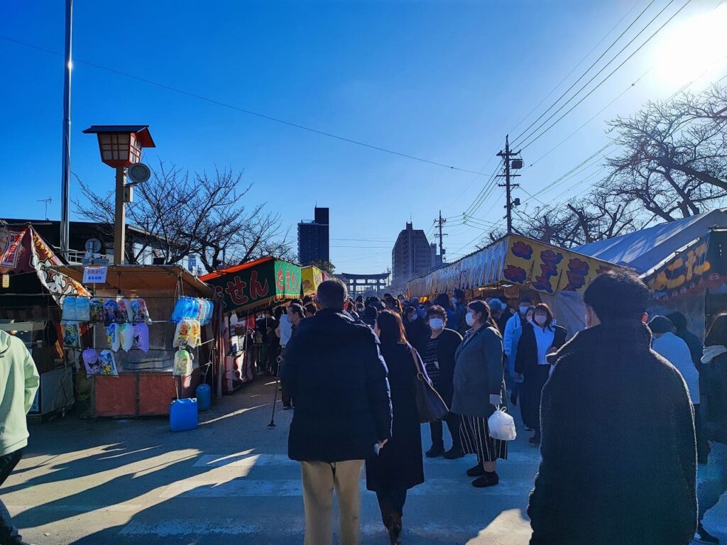 国府宮神社の参道に並ぶ初詣の屋台。夕方の時間帯で、参道沿いに複数の屋台が出店している。左側にはうどん・そばなどの飲食店の屋台があり、カラフルな看板や幟が掲げられている。多くの参拝者が冬服姿で屋台を眺めながら歩いている。背景には街灯や周辺のマンションが見え、初詣特有の賑やかな雰囲気が漂う。2024年1月2日撮影。