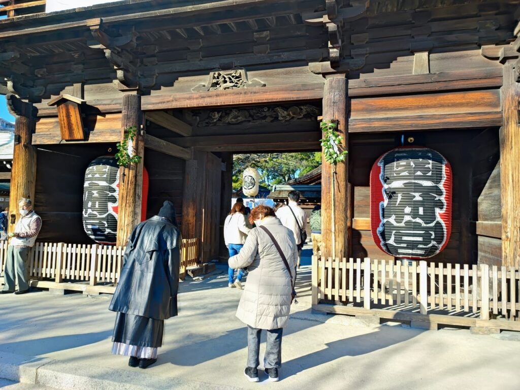 国府宮神社の楼門を間近から撮影。重厚な木造の楼門には精巧な彫刻が施され、左右に大きな白地と赤地の提灯が吊り下げられている。提灯には神社の紋章が描かれている。楼門の柱には松飾りが飾られ、新年の雰囲気を演出している。参拝客が楼門をくぐり抜ける様子が見られ、楼門の奥には拝殿や授与所が続いている。2024年1月2日撮影。