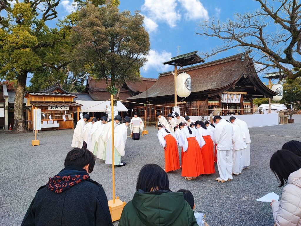 国府宮神社境内で執り行われる大祓式の神事。青空の下、拝殿前の広場に神職と巫女が整列している。神職は白い装束、巫女は白い上衣に朱色の袴姿で厳かに儀式を執り行っている。背景には重厚な本殿や拝殿の建物、大きな御神木が見え、左側には「おみくじ」と書かれた授与所がある。参拝者が後方から静かに見守る中、年末の穢れを祓う伝統的な神事が進められている。2025年12月31日撮影。