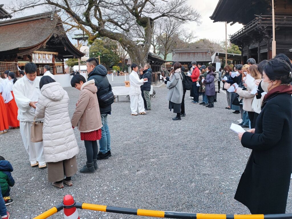 国府宮神社の大祓式で参拝者が切麻を体に振りかけた後の境内の様子。参道の地面には白い紙片(切麻)が散らばっている。左側には白装束に朱色の袴姿の巫女と参拝者が、右側には一般の参拝者が白い半紙を手に持ち、それぞれ切麻を体に振りかけて穢れを祓っている。背景には拝殿や大きな御神木、授与所などが見え、曇り空の下で厳かな雰囲気の中、年末の伝統行事が執り行われている。2025年12月31日撮影。
