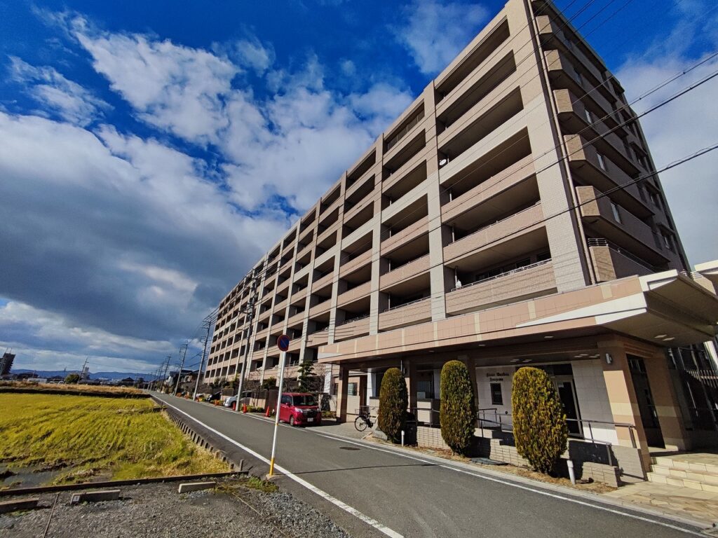 愛知県稲沢市に建つライオンズガーデン稲沢西棟の外観。道路に面したベージュ系の大規模マンションで、周辺は落ち着いた住環境が広がる不動産物件