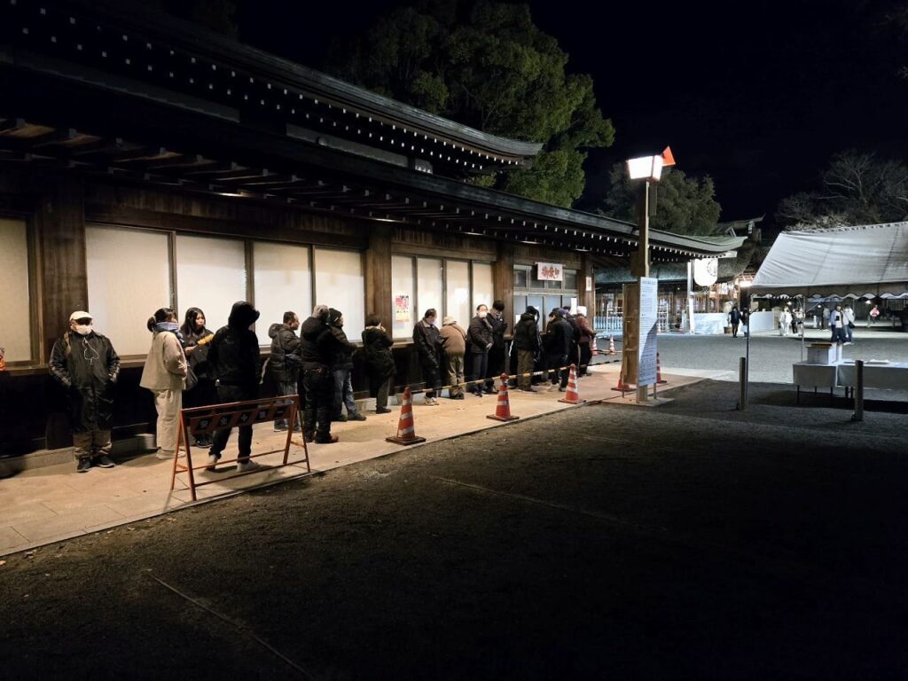 国府宮神社の交通安全祈願祭を待つ行列の様子。建物の前にコーンとバリケードが設置され、防寒着を着た参加者たちが一列に並んでいる。夜の境内で、背景に授与所のテントが見える。
