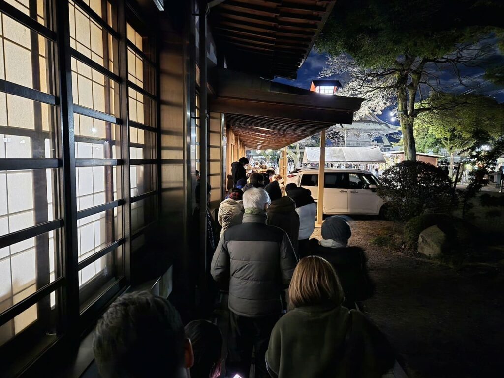 国府宮神社の廊下で交通安全祈願祭を待つ参加者たちの後ろ姿。障子から明かりが漏れる建物の横を歩いて進んでいる。防寒着を着た人々が列をなしている。