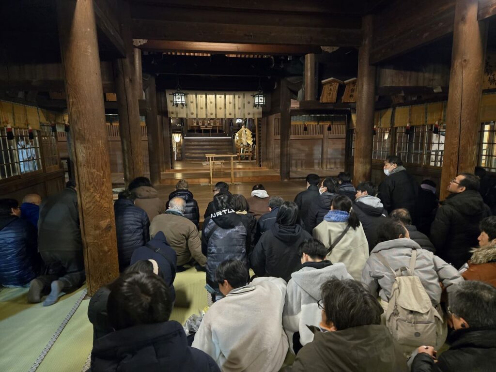 国府宮神社の拝殿内部の様子。太い木の柱が立ち並び、幣饌料と書かれた木の額が複数掛かっている。床には藁で編んだ円形の座具が置かれ、奥に本殿への階段が見える。参拝者が後ろから見守っている。