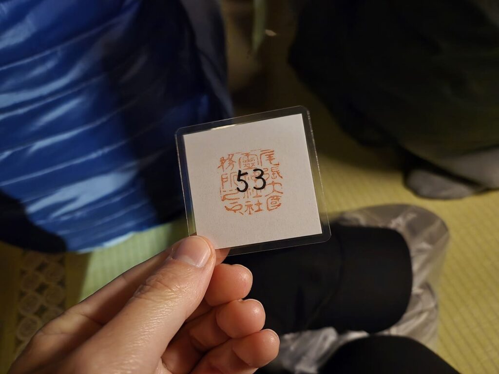国府宮神社の交通安全祈願祭で配布された番号札を手に持っている写真。白い紙に朱色の「尾張大國霊神社」の印が押され、中央に黒字で「53」と番号が書かれている。