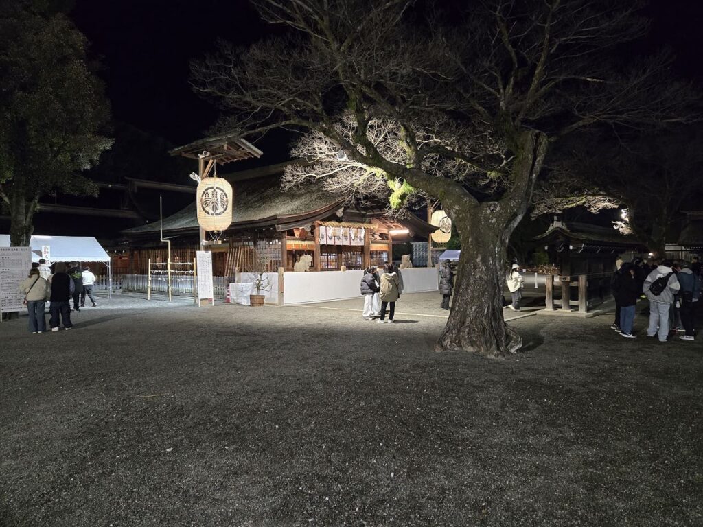 深夜の国府宮神社境内で授与品を求める参拝者たちの様子。「国府宮」と書かれた白いテントの下にお守りや絵馬が並び、多くの人が集まっている。提灯の明かりが境内を照らしている。