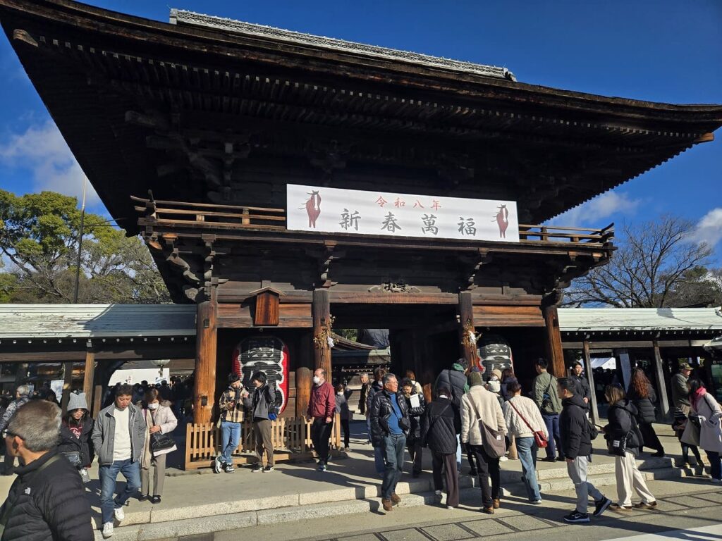 国府宮神社の楼門を正面から撮影した写真。「令和八年 新春萬福」と書かれた白い横断幕が掲げられ、木造の立派な楼門の下を多くの参拝者が行き来している。青空の下、初詣の賑わいを見せている。