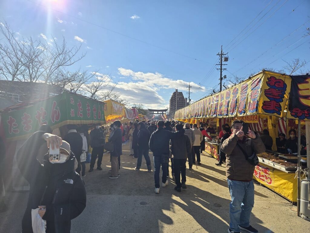 国府宮神社の参道に並ぶ屋台の様子。左側に緑色の「たません」の屋台、右側に黄色い「ねぎま」の屋台があり、多くの参拝者が歩いている。奥には鳥居が見え、青空と太陽の光が差し込んでいる。