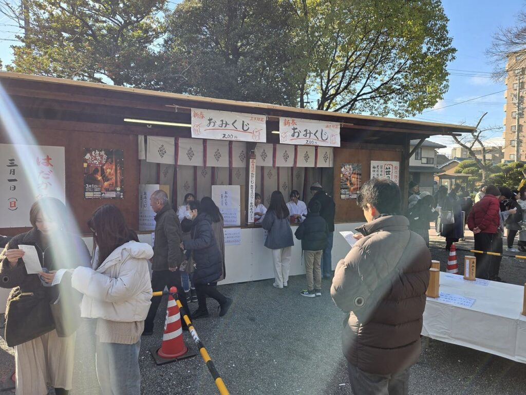 国府宮神社のおみくじ授与所の様子。木造の建物に「新春おみくじ 200円」と書かれた看板が掲げられ、白い装束を着た巫女さんが対応している。左側には「はだか祭 3月1日」のポスターが貼られている。