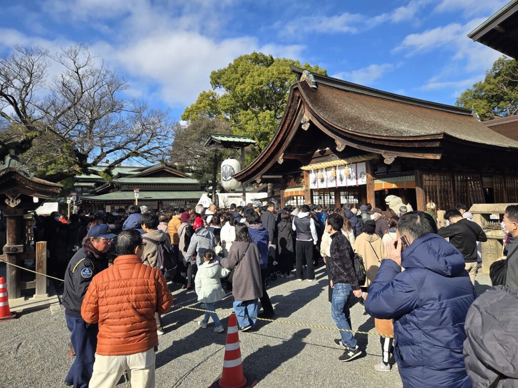 国府宮神社の拝殿前で参拝の順番を待つ大勢の参拝者たち。茅葺き屋根の拝殿が見え、境内は初詣客で混雑している。青空の下、防寒着を着た人々がロープに沿って並んでいる。