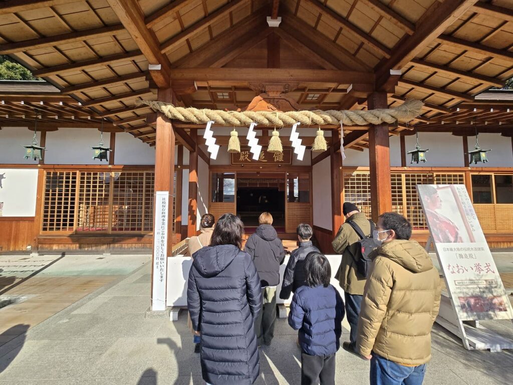 国府宮神社の儺追殿の正面から撮影した写真。しめ縄と紙垂が飾られた木造の建物の前で、数人の参拝者が手を合わせている。右側には「なおい拳式」の看板が立てられている。