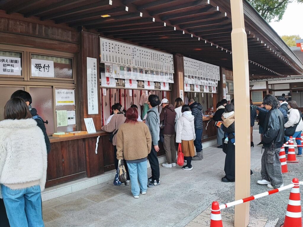 国府宮神社の授与所前に並ぶ参拝者たち。ジーンズやダウンジャケットなど普段着の人が多く、カジュアルな服装で参拝している様子が分かる。左側に「受付」の看板と祈祷申込の窓口がある。