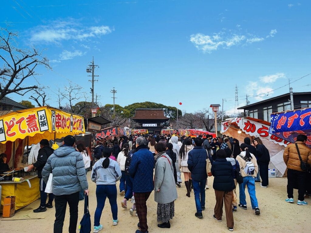 国府宮神社の参道に並ぶ屋台と参拝者。左側に「どて煮」「串かつ」の黄色い屋台、右側に「からあげ」の青い屋台があり、奥に楼門が見える。青空の下、多くの人で賑わっている。