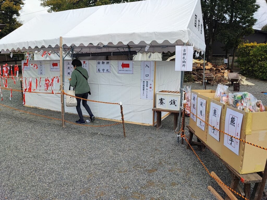 国府宮神社の古神札納所。白いテントの中に「古神札納所」の看板があり、入口には「なおいぎれ」「注連縄」「絵馬」「御守」「神札」の返納場所を示す案内が掲示されている。手前には「熊手」「破魔矢」の返納箱と賽銭箱が置かれている。