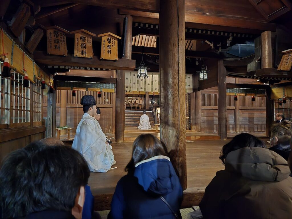 国府宮神社の拝殿で行われている交通安全祈願祭の様子。白い装束を着た神職が正座し、奥には本殿が見える。参拝者たちが後ろから見守っている。木造の荘厳な拝殿内部に幣饌料の額が掛かっている。