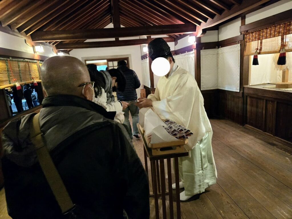 国府宮神社の交通安全祈願祭後、白い装束を着た神職が参加者に交通安全守を手渡している様子。参加者が一列に並び、順番にお守りを受け取っている。木造の建物内で行われている。