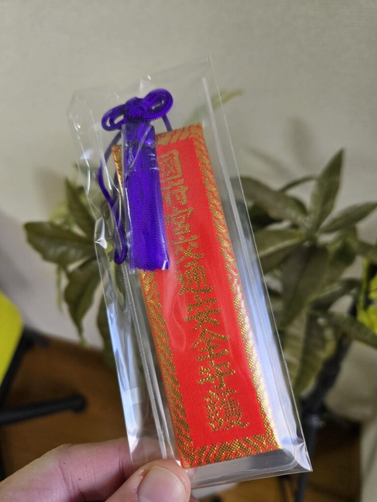国府宮神社の交通安全守を手に持っている写真。赤い布地に金色の刺繍で「国府宮交通安全守護」と書かれ、紫色の紐がついている。透明な袋に入った状態。