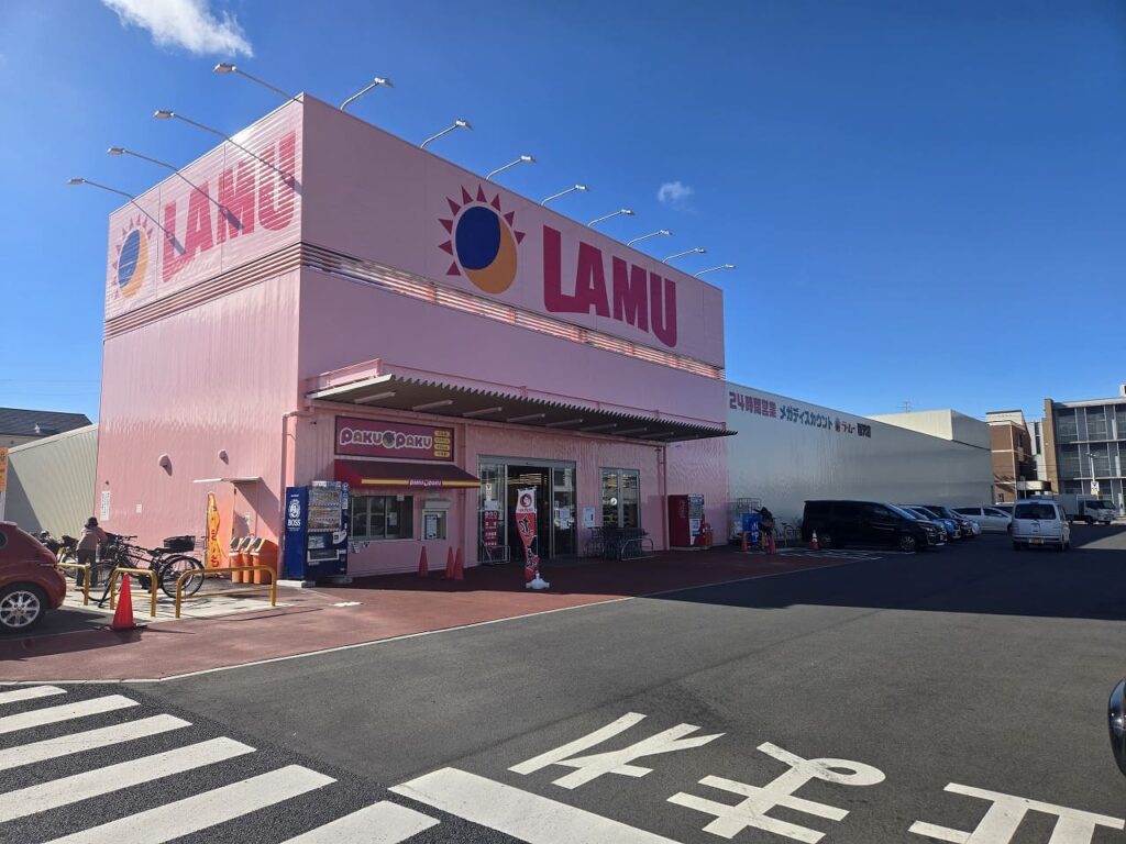 青空の下に広がるラ・ムー稲沢店の外観と、多くの車が停まっている広大な平面駐車場。店舗正面にはピンク色の大きなLAMUのロゴ看板が見える。