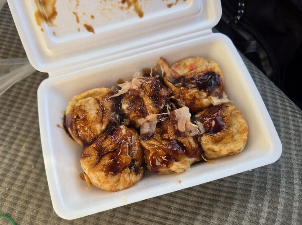 ラ・ムー稲沢店のPAKUPAKUで購入した焼きたてのたこ焼き。透明なパックに大粒のたこ焼きが6個入っており、ソースとマヨネーズ、かつお節がたっぷりとかかっている。