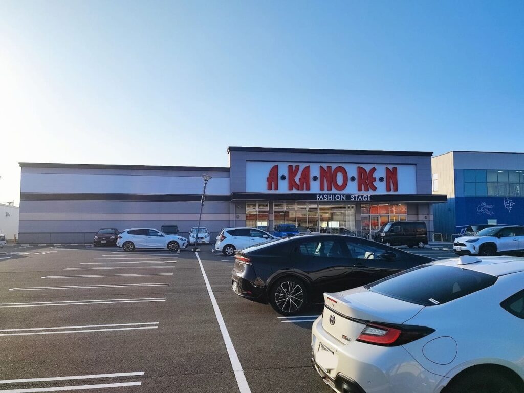 ニッケタウン稲沢内のA・KA・NO・RE・N(アカノレン)ファッションステージの外観。グレーの建物に赤い大きな看板が特徴的な衣料品店。広い駐車場には複数の車両が停車し、隣には他のテナントも見える