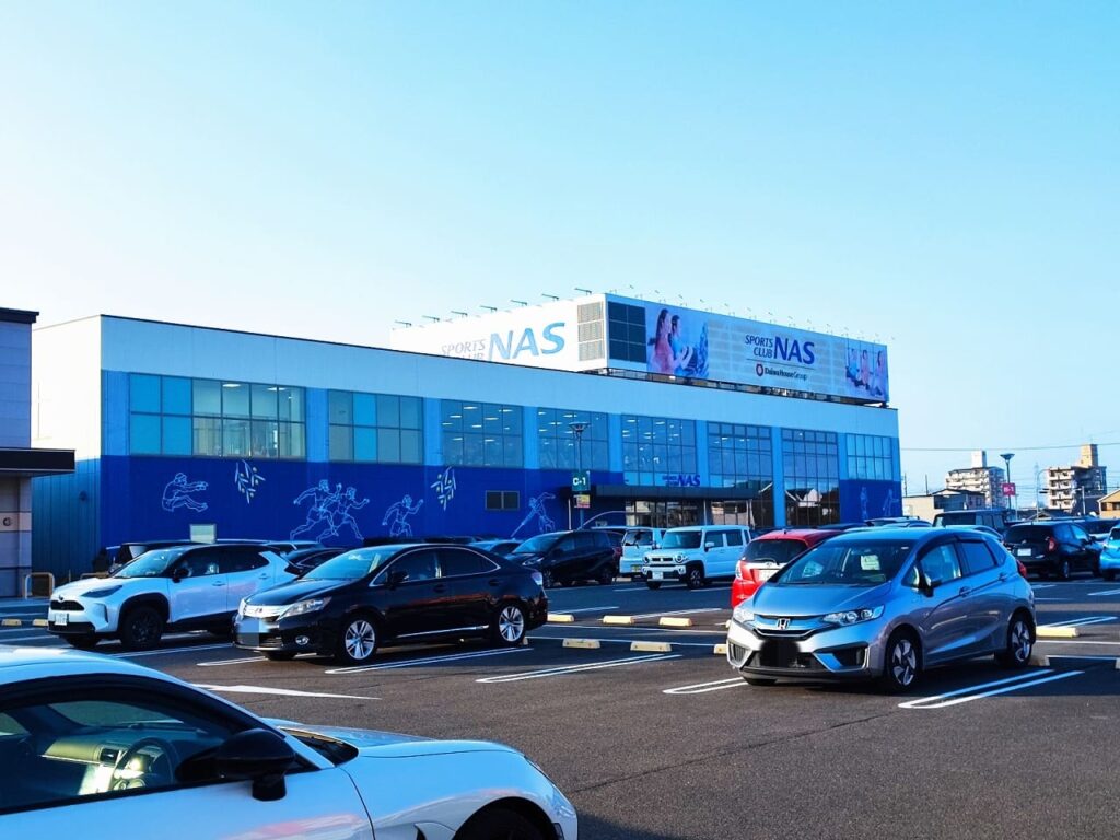 ニッケタウン稲沢のスポーツクラブNAS稲沢の外観。青い建物に大きなNASのロゴと白い文字が特徴的なスポーツ施設。広い駐車場には多数の車両が停車し、施設を利用する会員の姿も見える